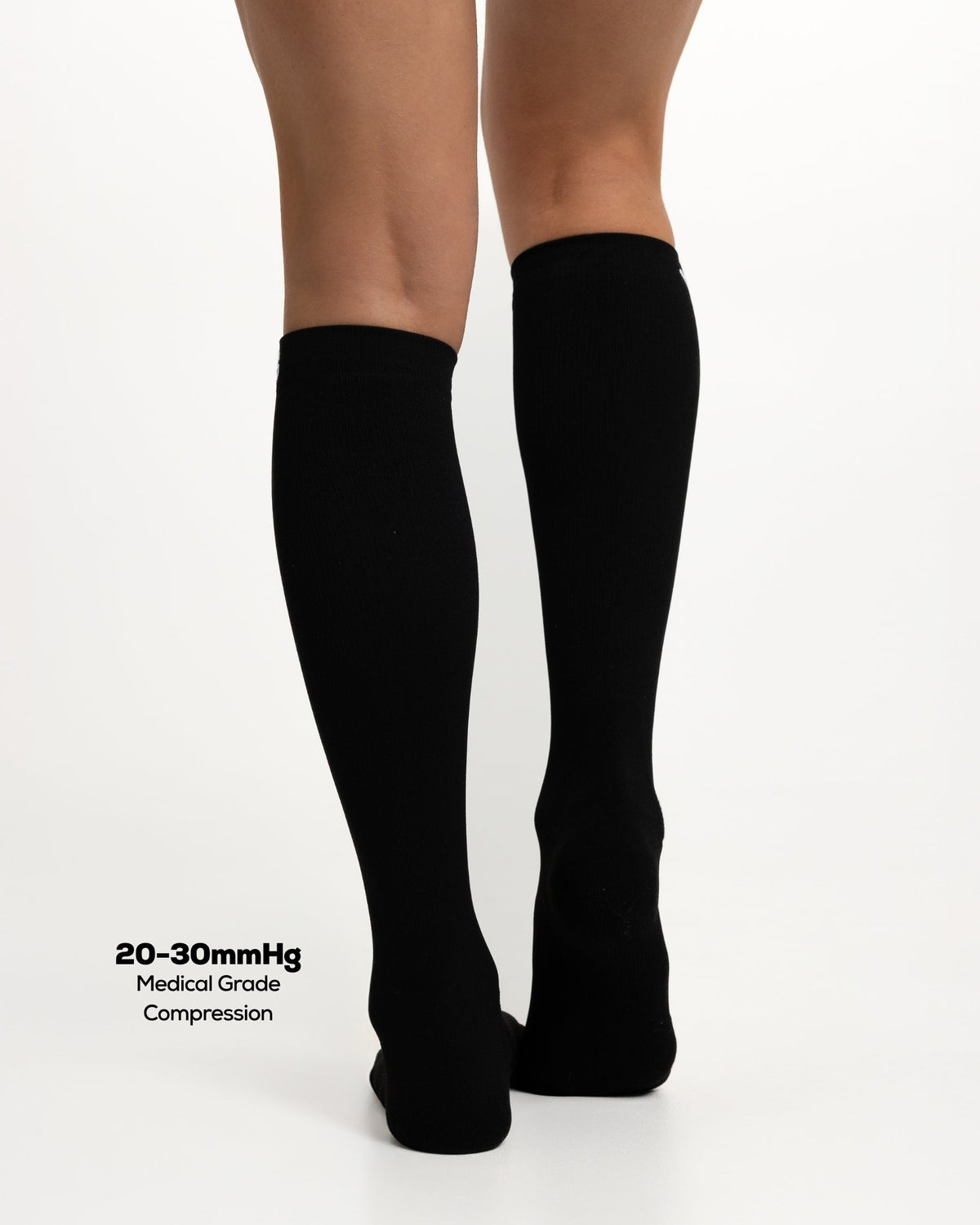 A person's legs from the back wearing a pair of black knee-high compression socks, shown against a plain white background with text reading '20-30mmHg Medical Grade Compression'.
