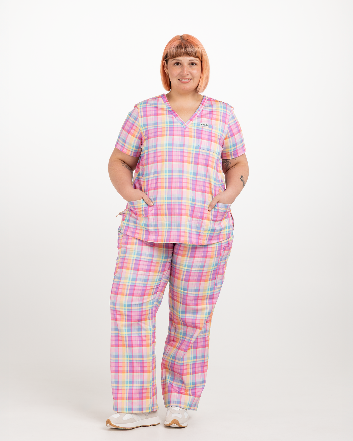 A woman modeling a matching two-piece scrub set with a pastel plaid pattern, featuring a short-sleeve V-neck top and coordinating pants, set against a plain white studio background.