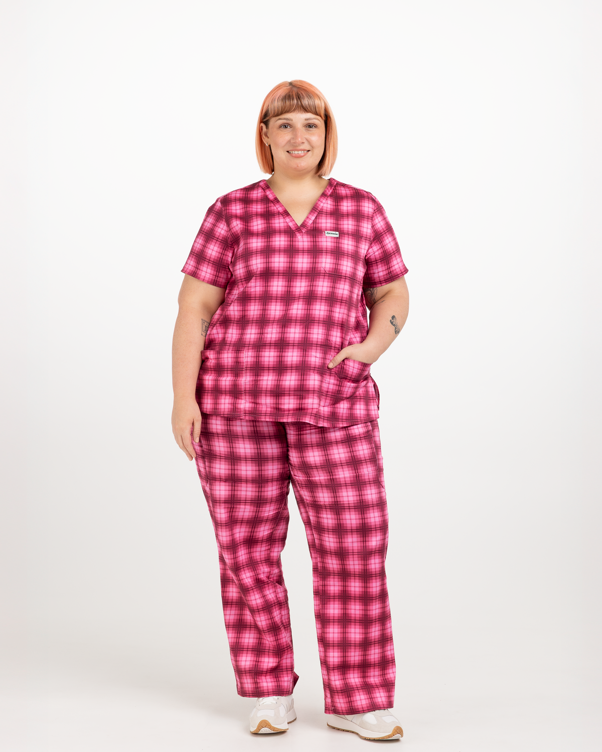 A woman modeling a matching pink and red plaid scrub set, consisting of a short-sleeve V-neck top and coordinating pants, against a plain white studio background.