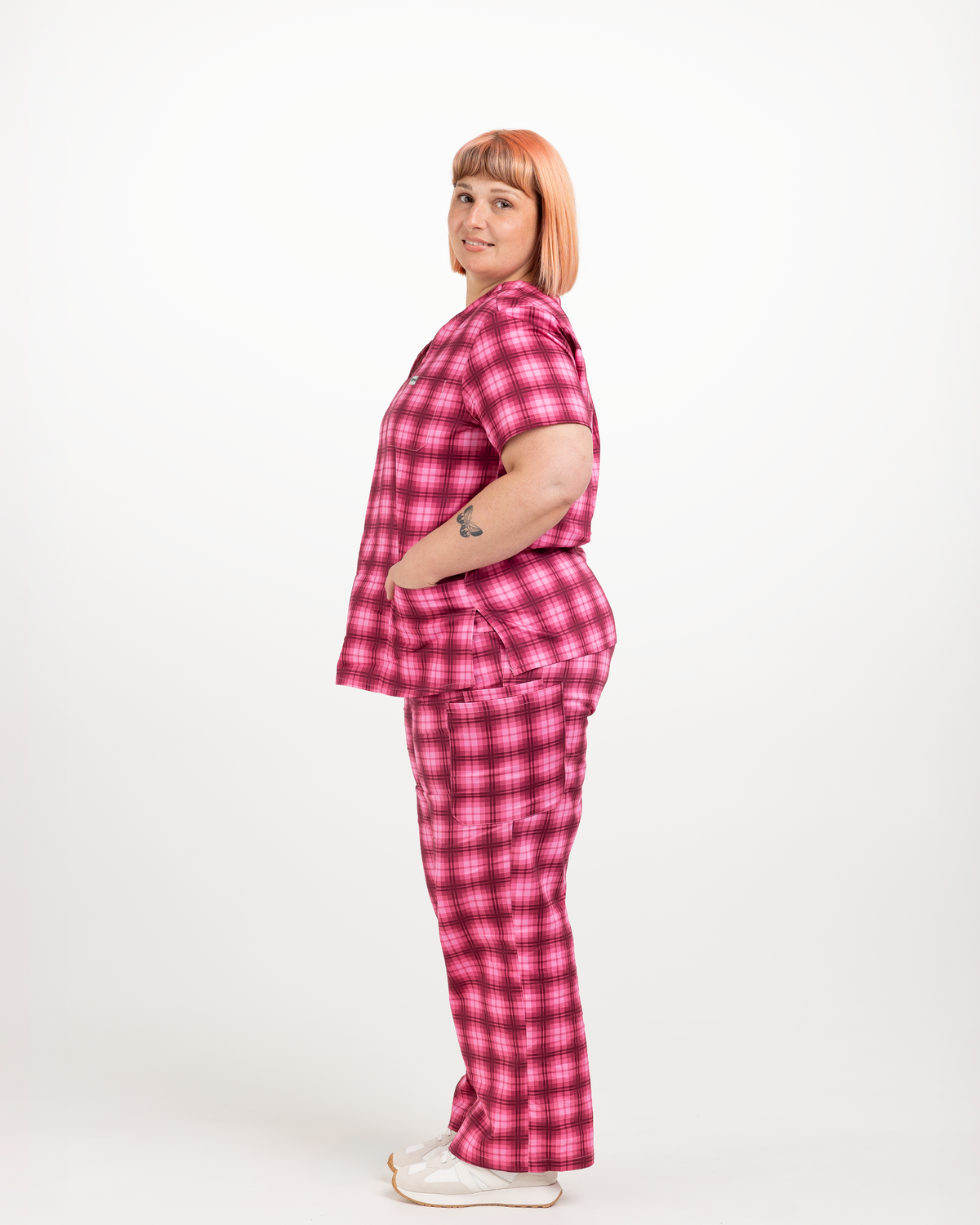 A woman with short pink hair stands in profile wearing a matching pink and red plaid short-sleeve top and pants set, paired with white slip-on shoes, against a plain white studio background.