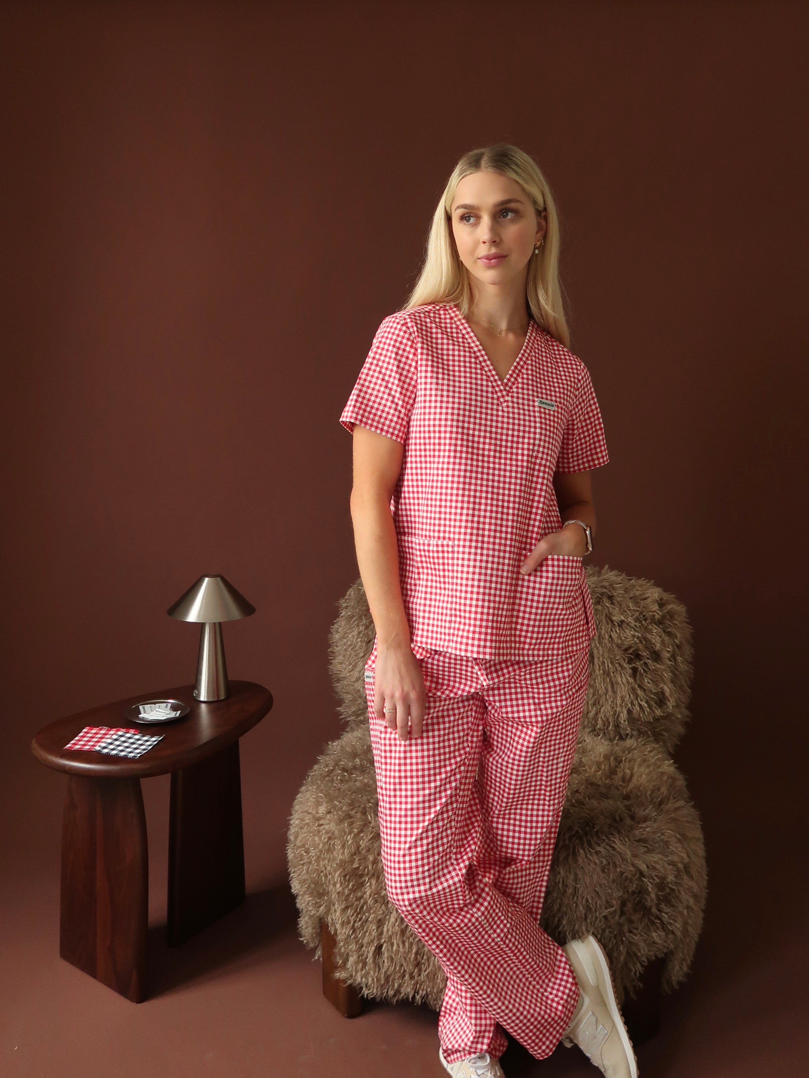 Model wearing Bennie Ruby Scrub Set in red checkered gingham print, styled with casual pose against brown backdrop.