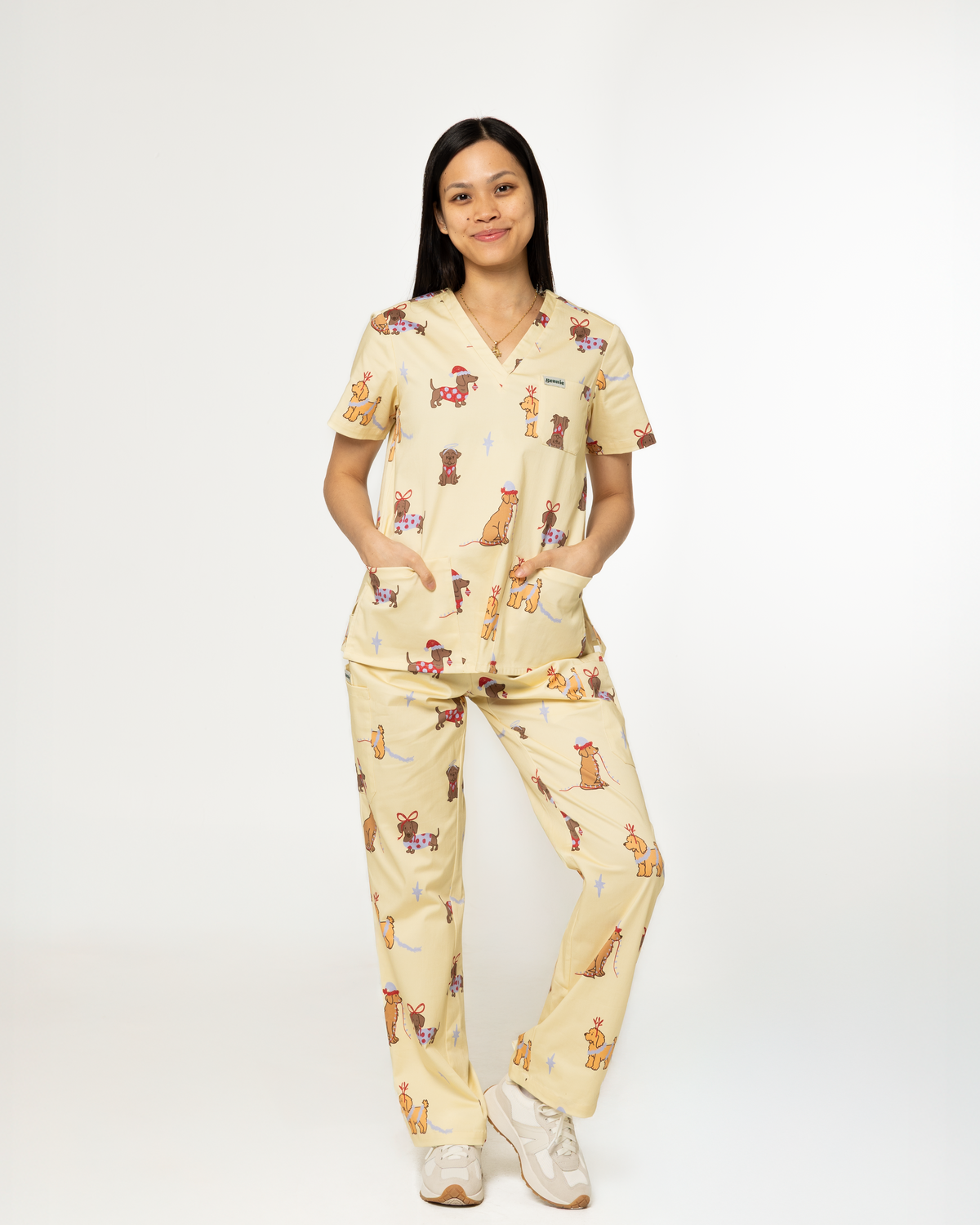 A woman modeling a matching medical scrub set with an all-over cartoon animal print on a light cream background, standing against a plain white studio background.